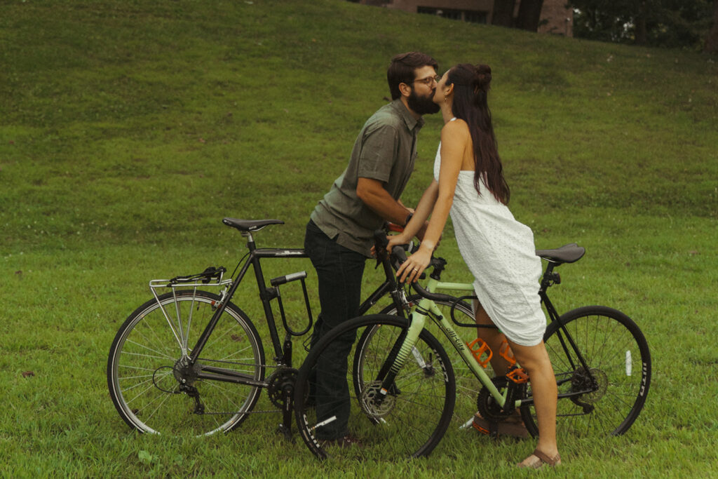 Couple kisses while standing beside matching bicycles in the grass, featuring playful 90s rom com romance and creative fun couple photoshoot ideas.