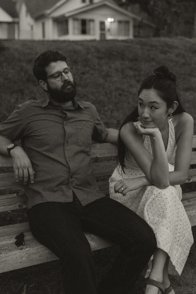 Black-and-white portrait of couple sitting on a bench making playful expressions, capturing quirky 90s rom com banter and fun couple photoshoot ideas.