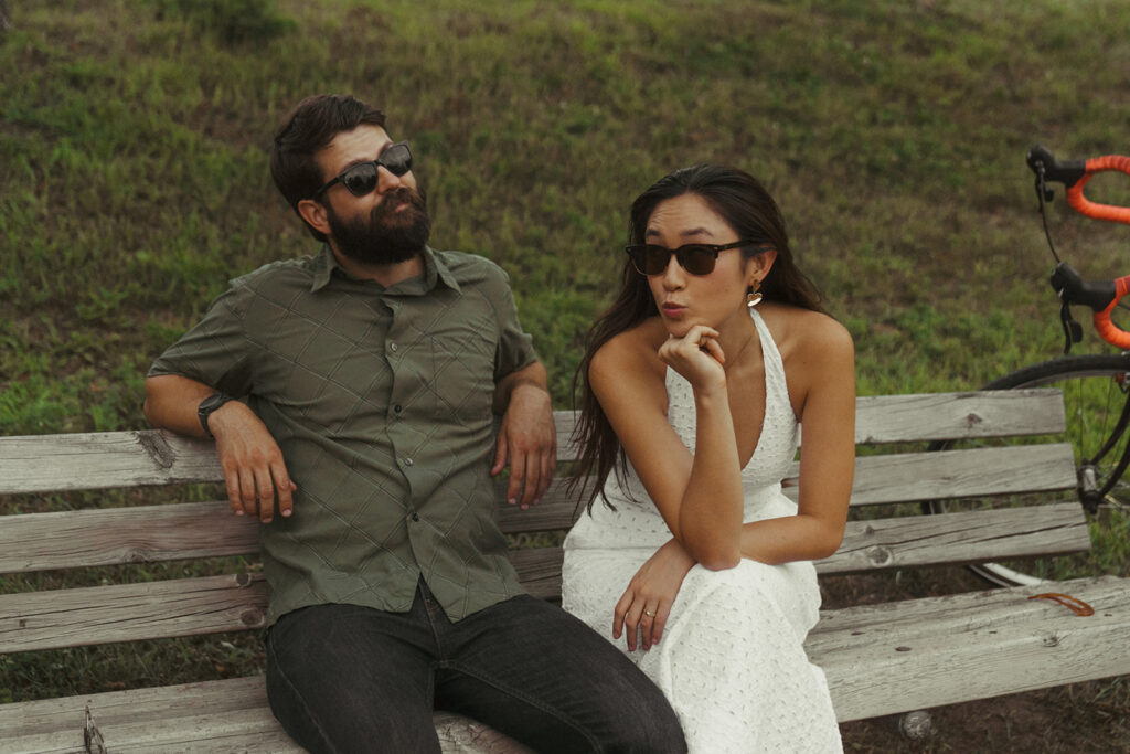 Couple wears sunglasses on a park bench beside their bike, capturing quirky 90s rom com vibes and fun couple photoshoot ideas full of personality.
