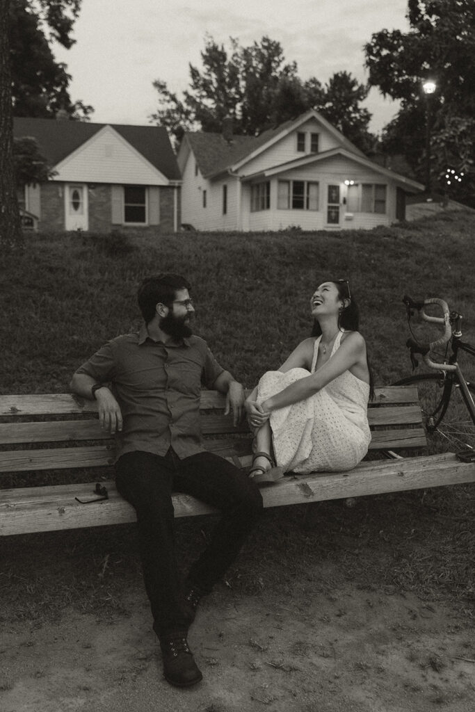 Black-and-white image of couple laughing together on a park bench with charming homes behind them, blending vintage engagement photos with nostalgic movie-scene energy.