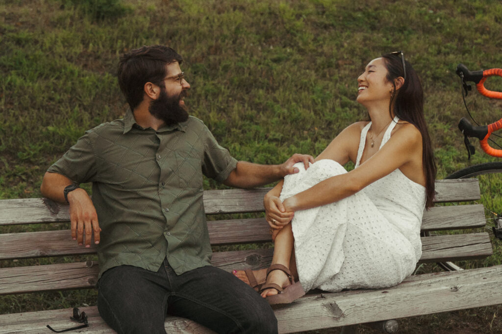 Couple laughs together on a weathered park bench beside their bikes, offering fun couple photoshoot ideas with warm Nancy Meyers aesthetic charm.