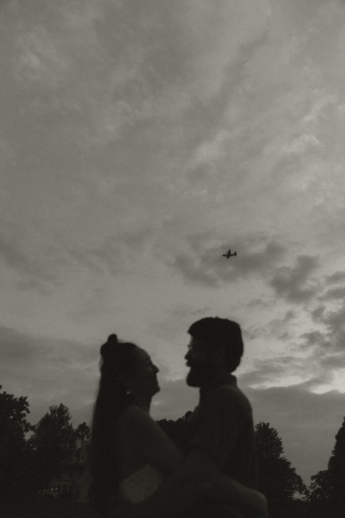Black-and-white silhouette of couple embracing at dusk with airplane overhead, creating cinematic vintage engagement photos with timeless 90s rom com mood.