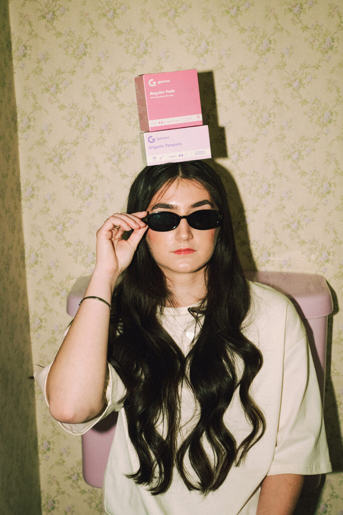 Woman in sunglasses seated in a vintage pink bathroom with Garnuu products balanced on her head for playful branding photos.