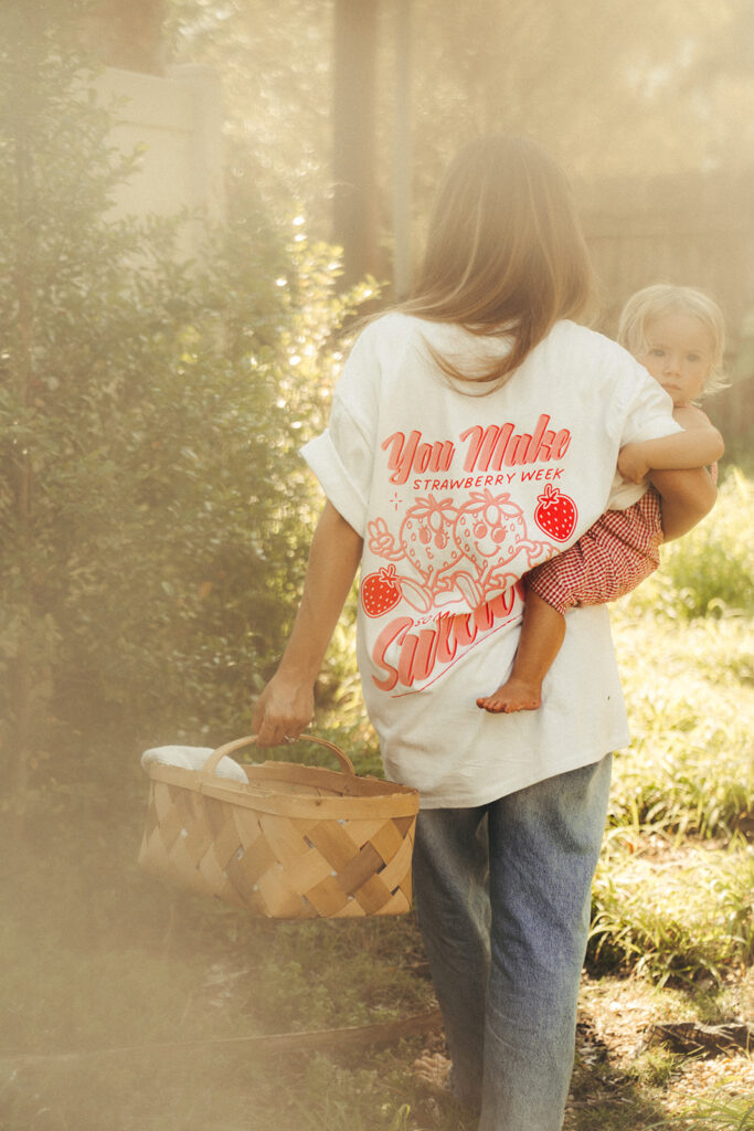 Mother carrying child with strawberry basket while wearing Garnuu shirt, heartfelt lifestyle branding photos.