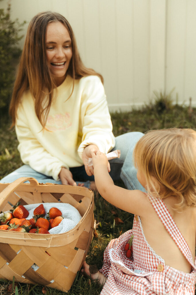 Woman handing toddler a Garnuu product beside strawberry basket, human lifestyle branding photos.