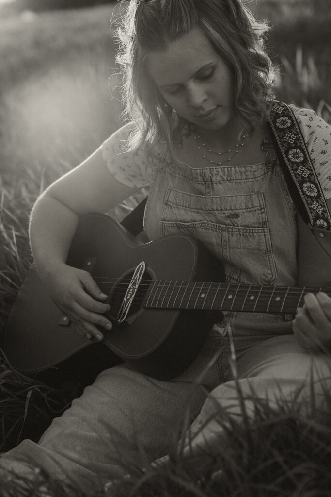 High schooler wearing denim overalls plays her guitar while sitting in Minnesota field