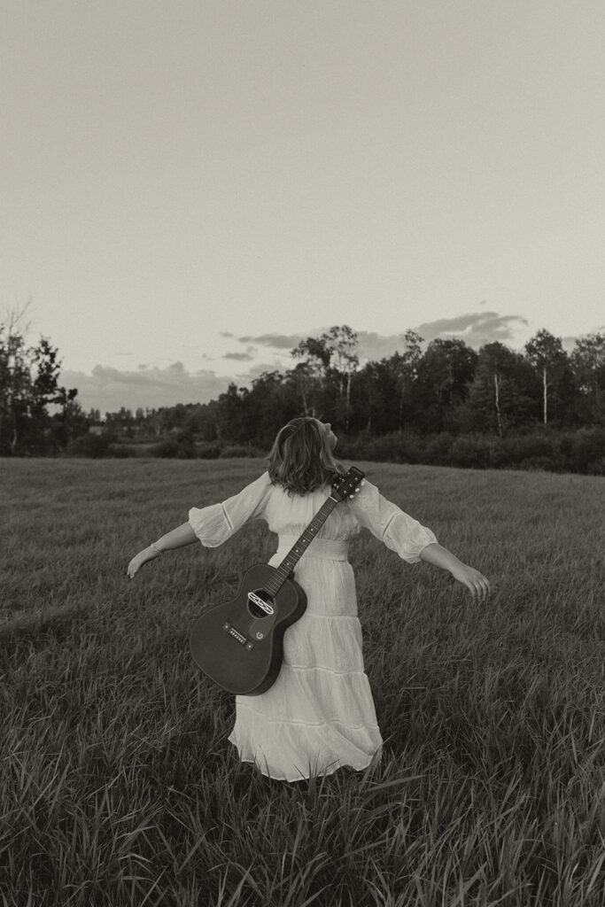 Girl in white dress twirls around with her guitar on her back in a Minnesota field