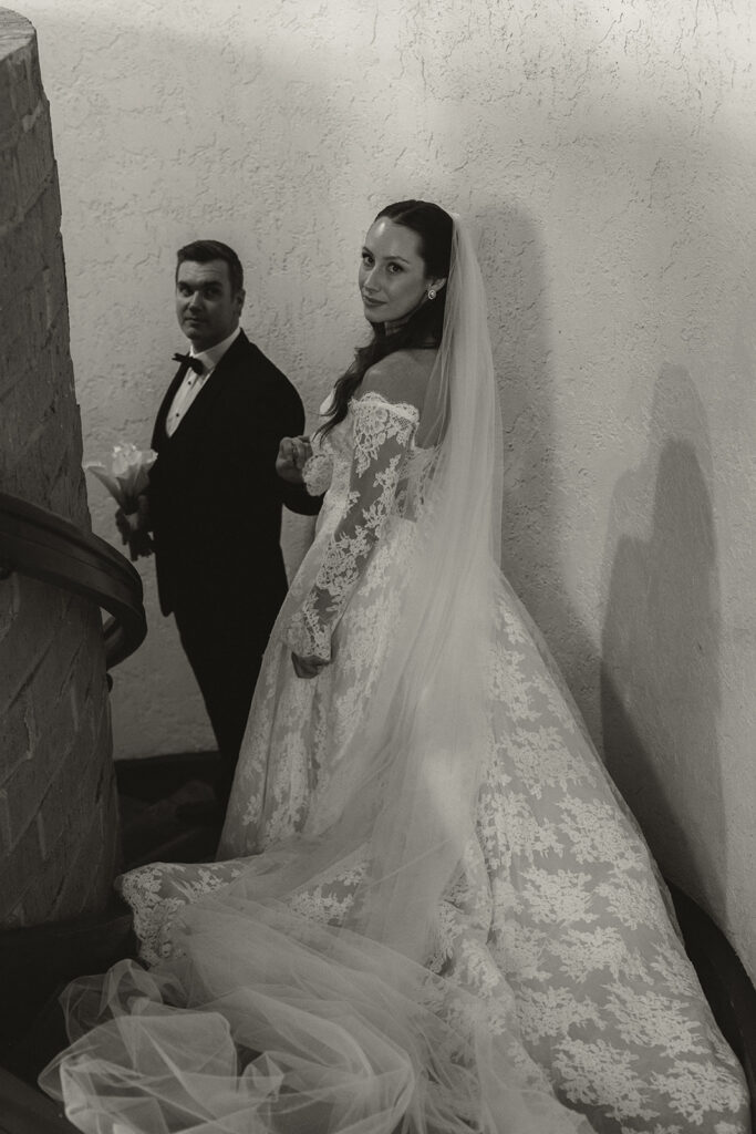 Bride looks back over her shoulder as groom looks back at her while they make their way down grand staircase at Edward Anne Estate