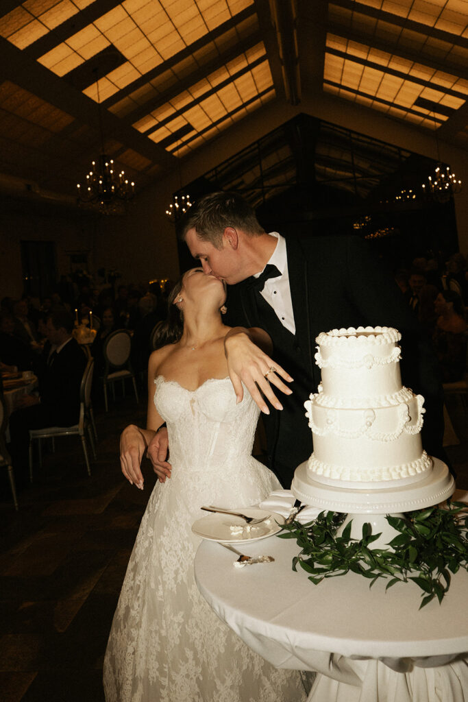 Bride shows off her ring and kisses her groom next to their two-tiered vintage wedding cake