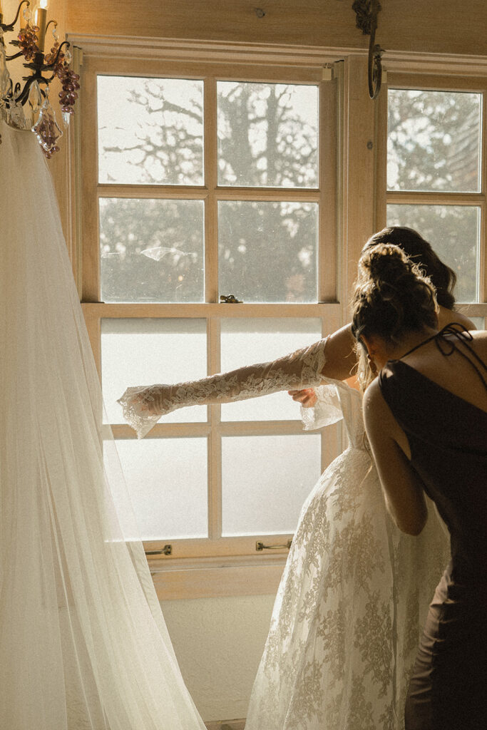 Bride stands in the window light of historic wedding venue while her mom helps button up her dress