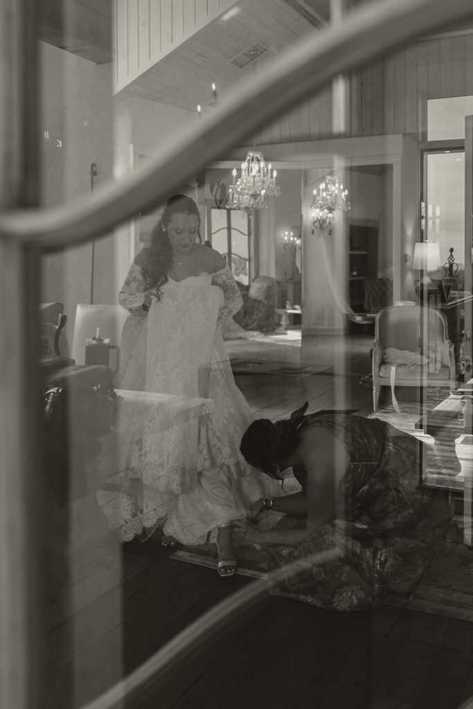 View through the glass door pane as bridesmaid helps bride clasp her shoes in Edward Anne Estate bridal suite
