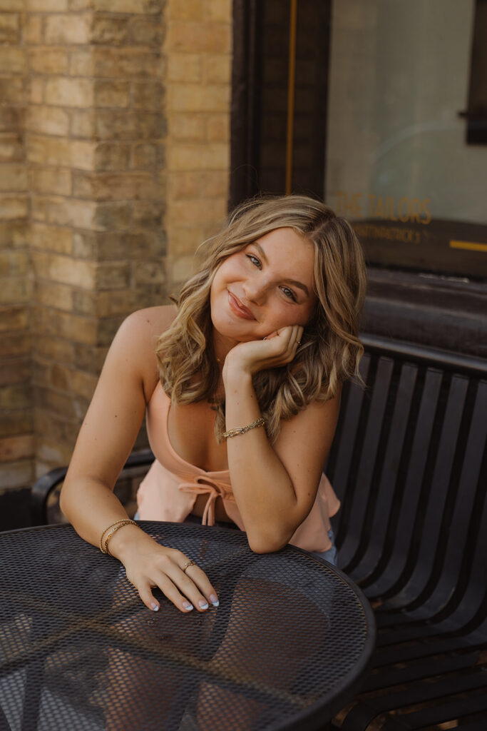Girl in pink tank top and denim skirt rests her head in her hands and soft smiles while sitting at a black bistro table
