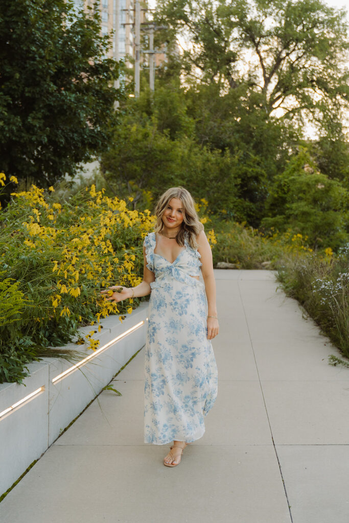Girl in blue and white floral sundress walks along city sidewalk framed by black eyed Susans