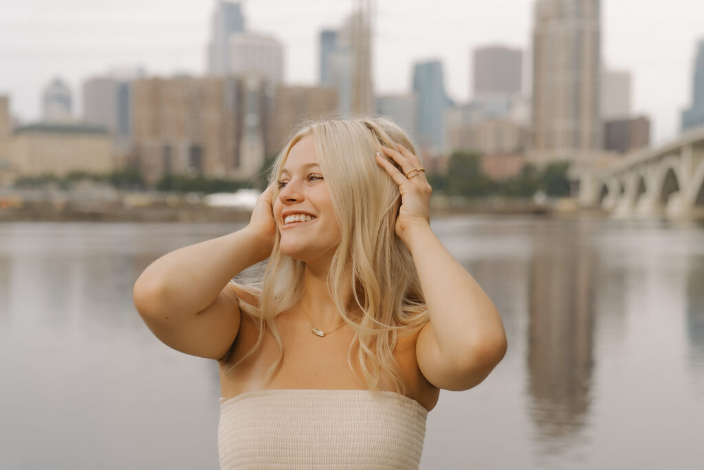 Blonde girl with gold rings looks off into the distance during her senior photos, Minneapolis skyline a blur in the background