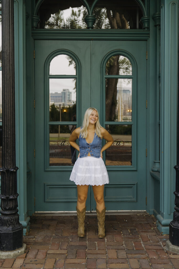 Young girl in cowboy boots, white skirt, and denim vest poses in front of dark green set of doors