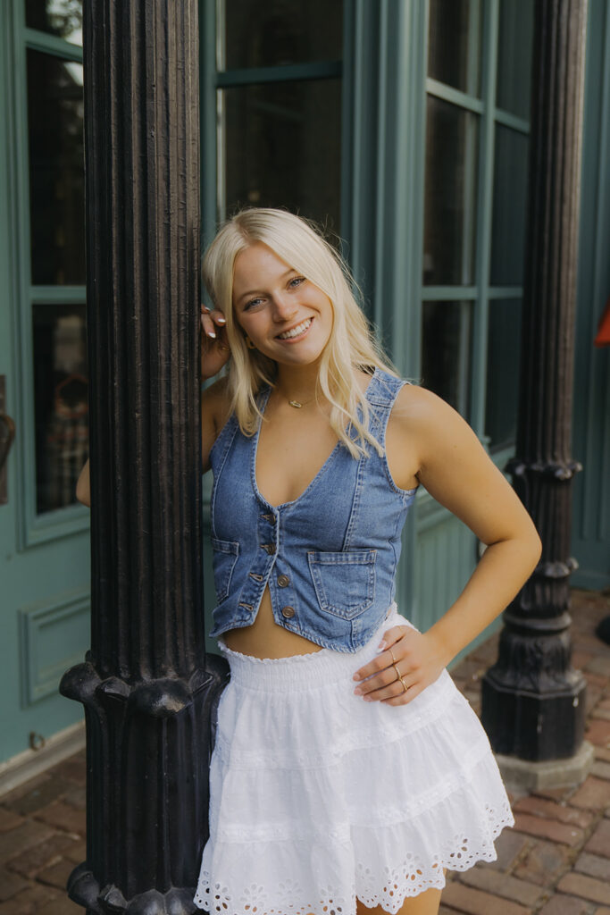 Girl in white skirt and denim vest leans her head against a lamppost in Minnesota senior photos