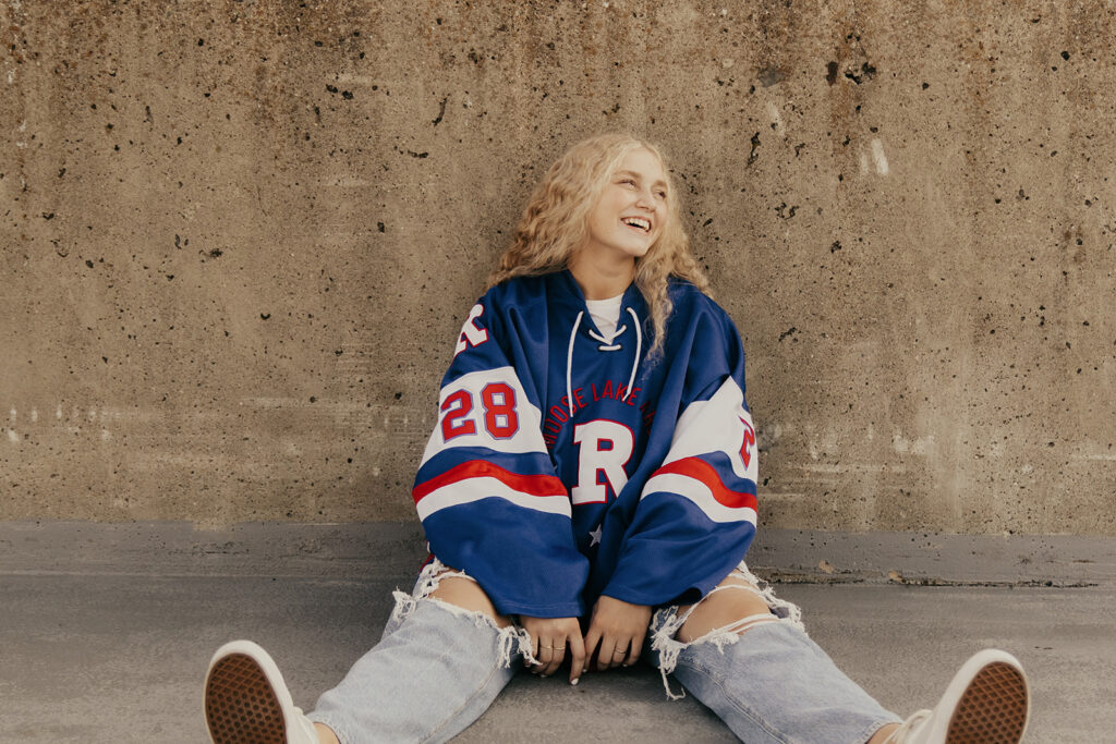 High school senior sits against concrete wall in her hockey jersey during senior photos