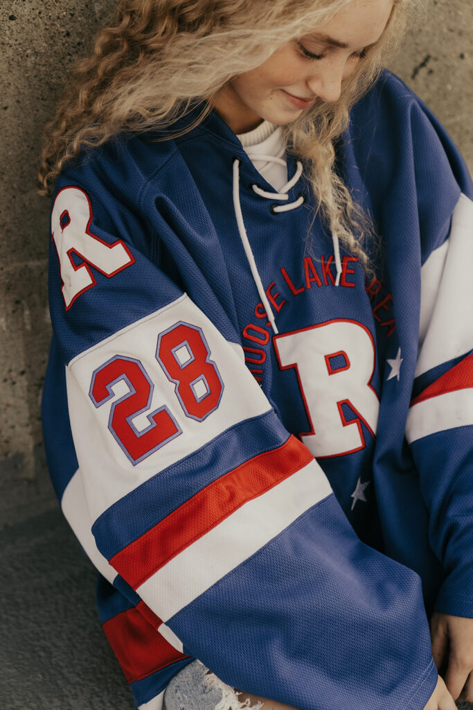 Girl with blonde, curly hair looks down at her Minnesota hockey jersey during senior photos