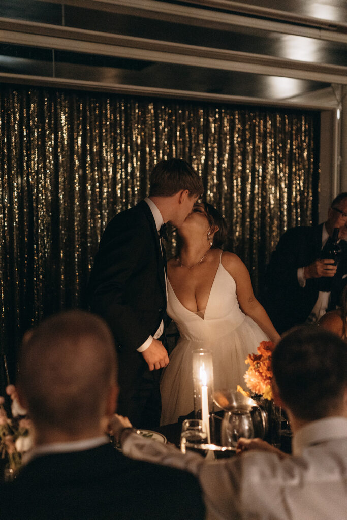 Bride and groom kiss over candlelight as wedding guests look on at The Commodore in Saint Paul, Minnesota