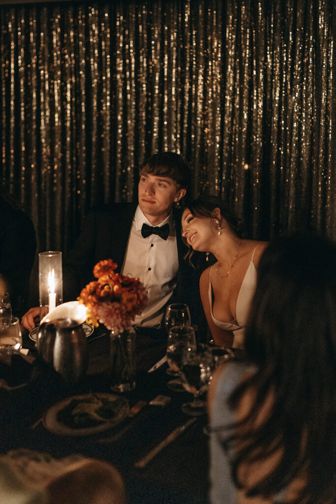 Bride leans her head on groom's shoulder while they listen to speeches over candlelight at The Commodore in Saint Paul, Minnesota