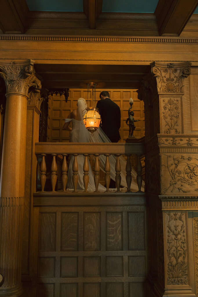 Bride and groom stand hand in hand on balcony at Van Dusen Mansion, surrounded by grand architecture