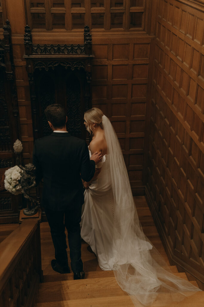 Groom's hand drifts to bride's shoulder to steady her as they make their way down staircase of Van Dusen Mansion