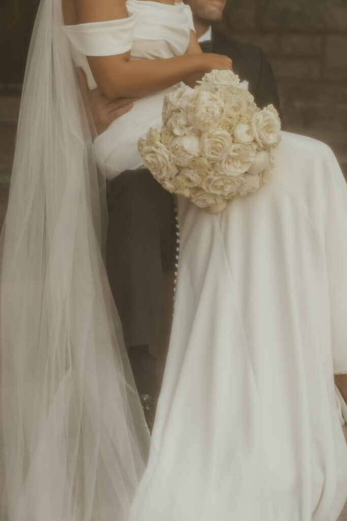 Bride sits on groom's lap while holding a bouquet of white roses