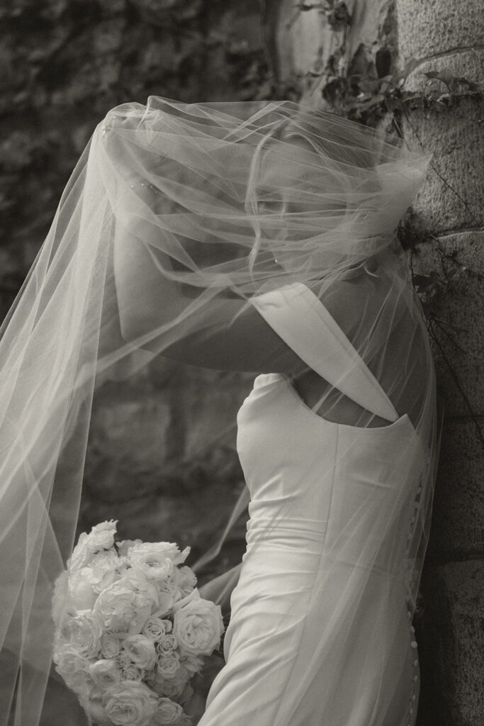 Bride leans against stone pillar at Van Dusen Mansion while gazing through her veil at the camera