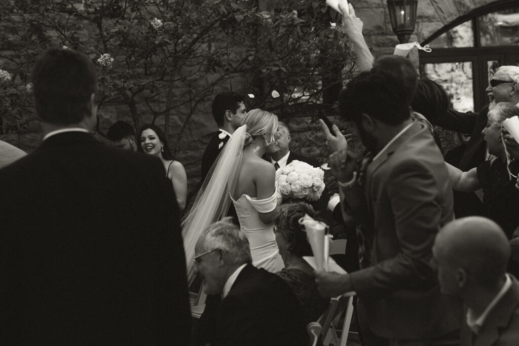 Guest toss white rose petals as newlyweds make their way down the aisle of courtyard at historic wedding venue in Twin Cities